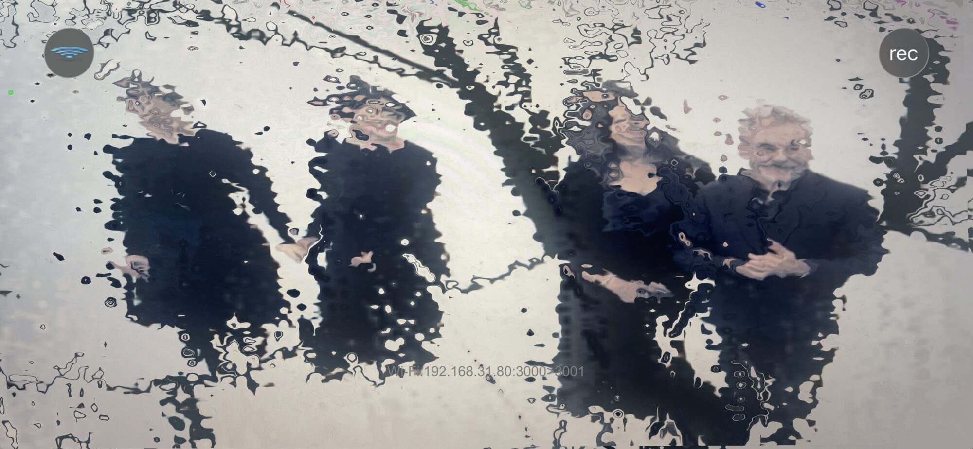 A phone held in the audience showing the stage through the app's rain
            effect, the actors dissolving behind running water
